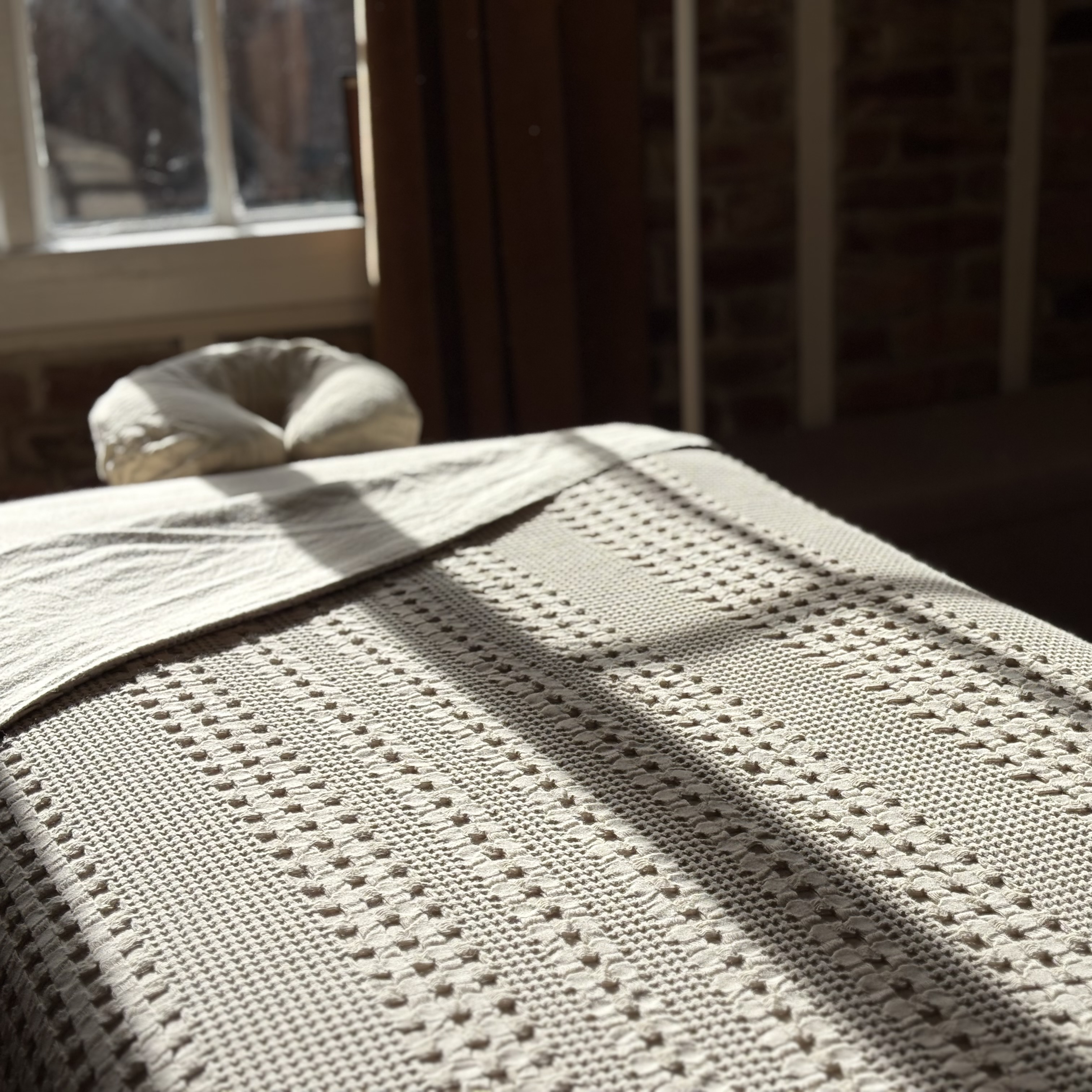 A sunlit massage table with textured cream linens beside a window